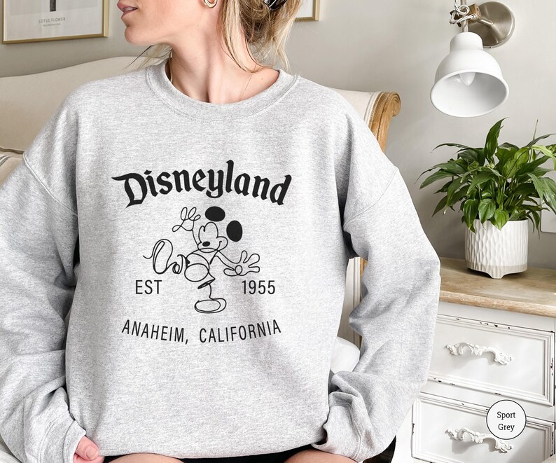 Puede incluir: Una sudadera gris claro con el logotipo de Disneyland en negro, con Mickey Mouse. El texto incluye "EST 1955" y "ANAHEIM, CALIFORNIA". La sudadera es de color gris deportivo.