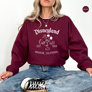 Puede incluir: Sudadera burdeos de Disneyland con letras blancas y un gr&aacute;fico de Mickey Mouse. El texto incluye "Disneyland", "EST 1955" y "Anaheim, California". La sudadera tiene cuello redondo y mangas largas.