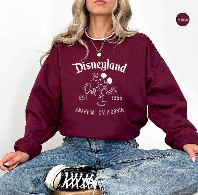 Puede incluir: Sudadera burdeos de Disneyland con letras blancas y un gr&aacute;fico de Mickey Mouse. El texto incluye "Disneyland", "EST 1955" y "Anaheim, California". La sudadera tiene cuello redondo y mangas largas.