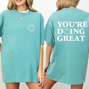 Puede incluir: Camiseta verde mar con una carita sonriente blanca en la parte delantera y la frase "YOU'RE DOING GREAT" en la espalda. La camiseta tiene mangas cortas y un corte holgado. El texto y la carita sonriente est&aacute;n en negrita, sin serifa.
