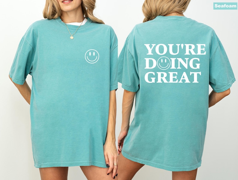 Puede incluir: Camiseta verde mar con una carita sonriente blanca en la parte delantera y la frase "YOU'RE DOING GREAT" en la espalda. La camiseta tiene mangas cortas y un corte holgado. El texto y la carita sonriente est&aacute;n en negrita, sin serifa.