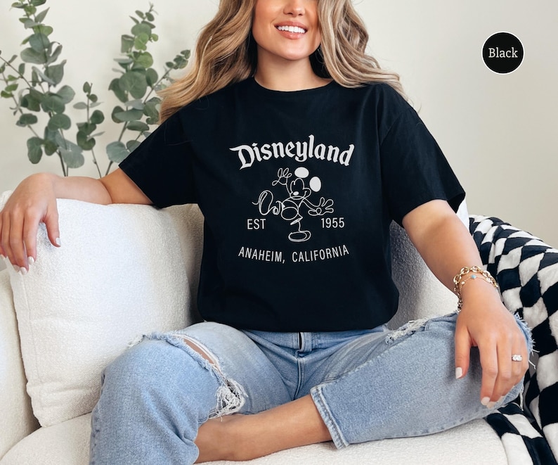 Puede incluir: Camiseta negra con el logo de Disneyland en blanco, con Mickey Mouse y el texto "EST 1955 Anaheim, California." La camiseta es de estilo cuello redondo cl&aacute;sico.