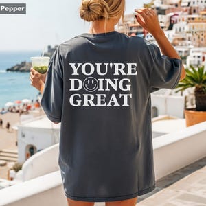 Puede incluir: Camiseta holgada color pimienta con el texto blanco "YOU'RE DOING GREAT" y una carita sonriente. La camiseta tiene un corte relajado, ideal para uso diario. La persona sostiene una bebida.