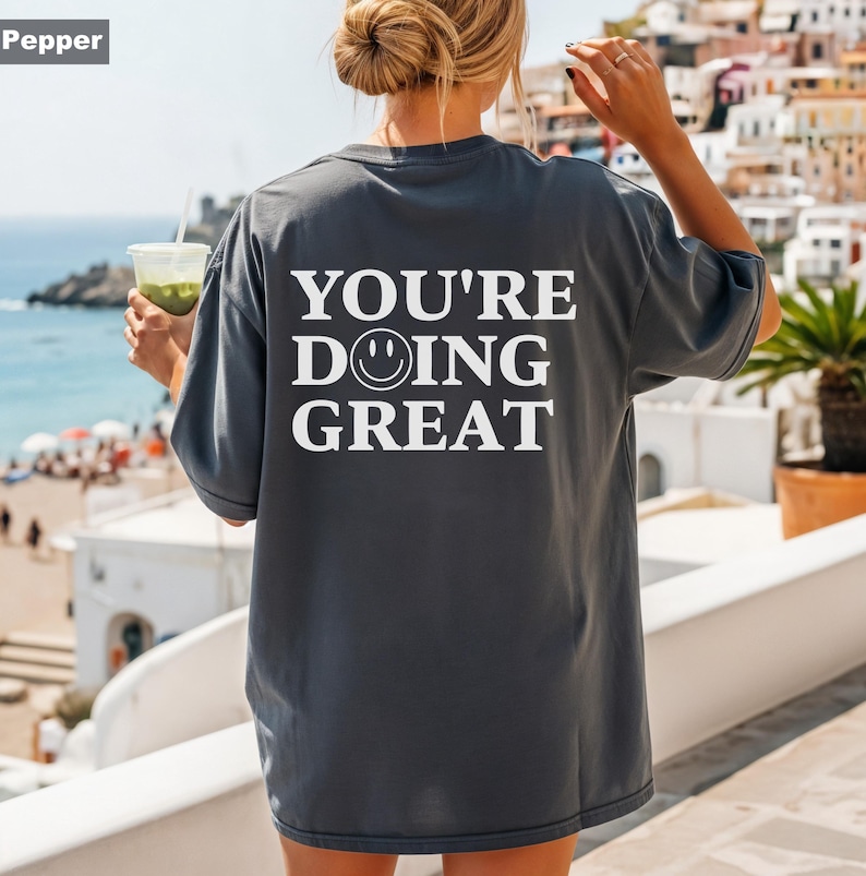 Puede incluir: Camiseta holgada color pimienta con el texto blanco "YOU'RE DOING GREAT" y una carita sonriente. La camiseta tiene un corte relajado, ideal para uso diario. La persona sostiene una bebida.