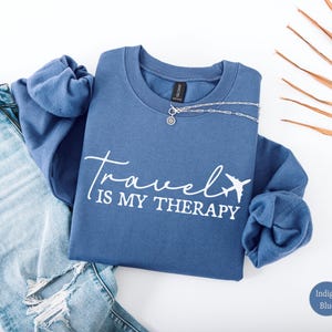May include: Indigo blue sweatshirt with the text "Travel is my Therapy" in white script, featuring a small airplane graphic. A silver necklace with a pendant is draped across the sweatshirt. Light blue denim jeans are visible on the left.