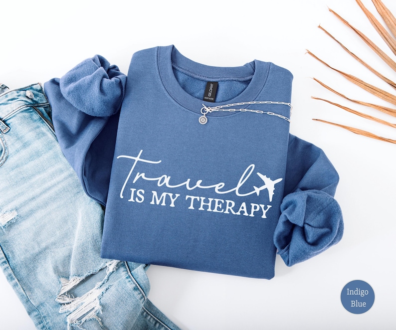 May include: Indigo blue sweatshirt with the text "Travel is my Therapy" in white script, featuring a small airplane graphic. A silver necklace with a pendant is draped across the sweatshirt. Light blue denim jeans are visible on the left.