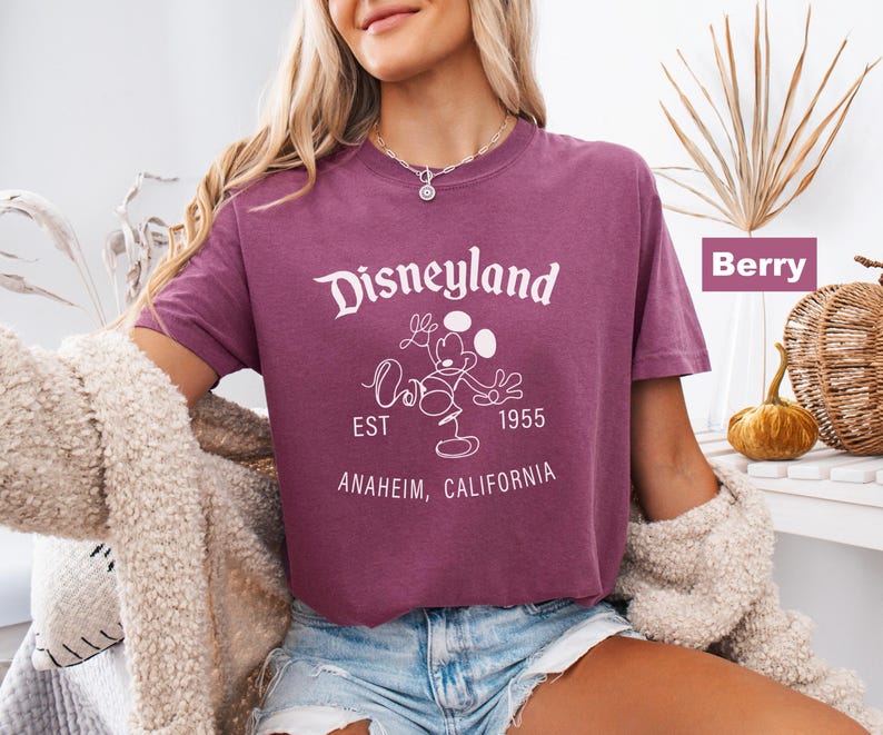 Puede incluir: Camiseta color bayas con el logo de Disneyland en blanco, con Mickey Mouse y el texto "EST 1955 Anaheim, California." La camiseta se combina con pantalones cortos de mezclilla.