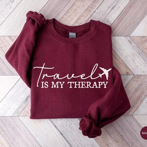 May include: A maroon sweatshirt featuring the white text "Travel is my Therapy" and a white airplane graphic. The garment is displayed on a light wood-effect surface. The sleeves are folded. The colour is described as maroon.