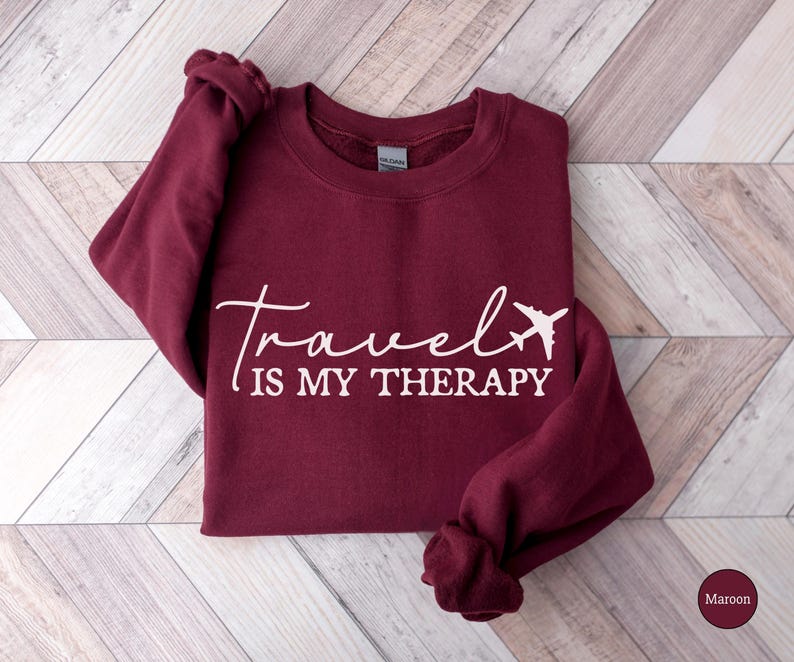May include: A maroon sweatshirt featuring the white text "Travel is my Therapy" and a white airplane graphic. The garment is displayed on a light wood-effect surface. The sleeves are folded. The colour is described as maroon.