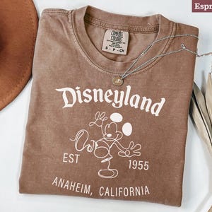 Puede incluir: Camiseta color espresso con el logo de Disneyland en blanco, con Mickey Mouse. La camiseta incluye el texto "EST 1955 ANAHEIM, CALIFORNIA". Un collar plateado con un colgante est&aacute; sobre la camiseta.
