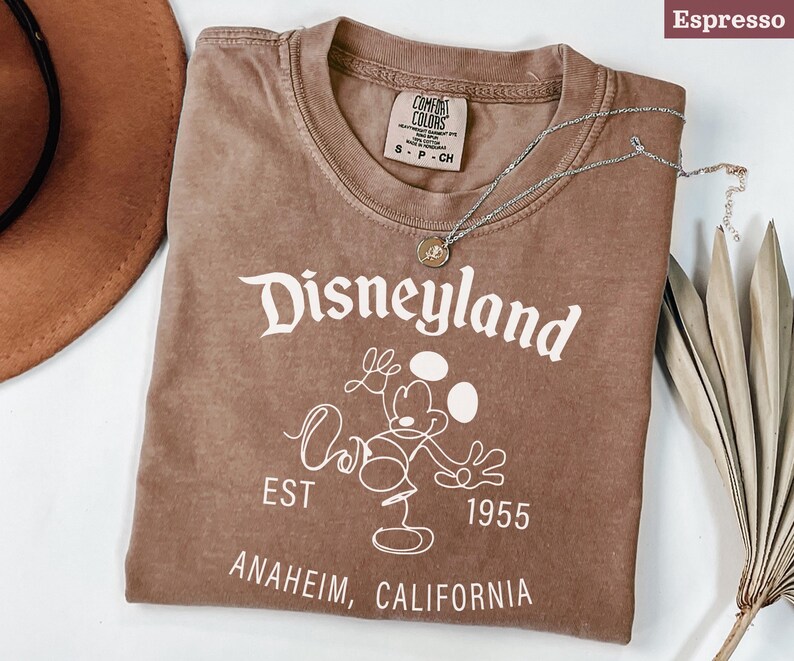 Puede incluir: Camiseta color espresso con el logo de Disneyland en blanco, con Mickey Mouse. La camiseta incluye el texto "EST 1955 ANAHEIM, CALIFORNIA". Un collar plateado con un colgante est&aacute; sobre la camiseta.