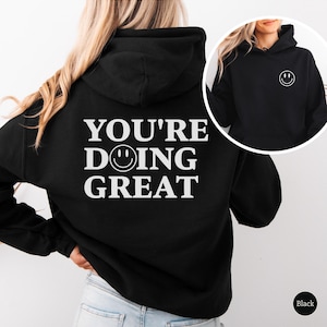 Puede incluir: Sudadera con capucha negra con el texto "YOU'RE DOING GREAT" y una carita sonriente. La misma carita sonriente tambi&eacute;n est&aacute; en la parte delantera. Estilo casual.