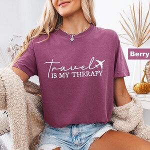 May include: Berry-coloured t-shirt with the text "Travel is my therapy" in white script and a small airplane graphic. The shirt is a short-sleeved crew neck style.