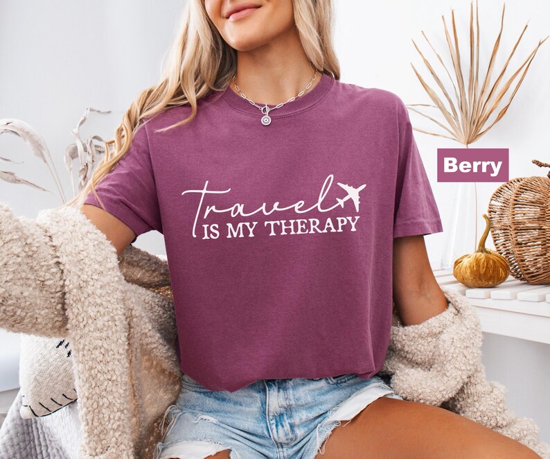 May include: Berry-coloured t-shirt with the text "Travel is my therapy" in white script and a small airplane graphic. The shirt is a short-sleeved crew neck style.