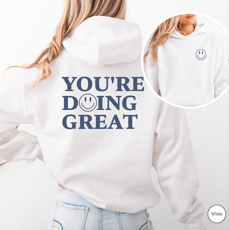 Puede incluir: Sudadera con capucha blanca con el texto "YOU'RE DOING GREAT" en azul marino en la espalda. Una peque&ntilde;a carita sonriente azul marino est&aacute; encima de la palabra "DOING". Una imagen m&aacute;s peque&ntilde;a de la sudadera con capucha con una carita sonriente est&aacute; en la esquina superior derecha.