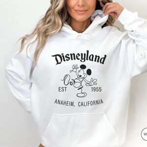 Puede incluir: Sudadera con capucha blanca con el logotipo de Disneyland, Mickey Mouse, el texto "EST 1955" y "Anaheim, California" en negro. La sudadera tiene un bolsillo delantero y una capucha con cord&oacute;n.