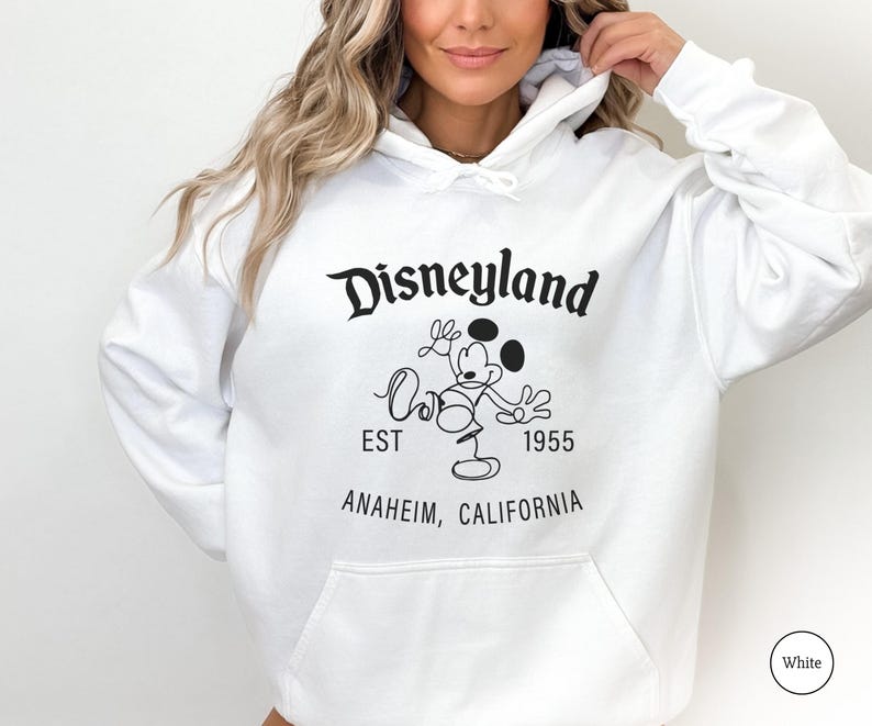 Puede incluir: Sudadera con capucha blanca con el logotipo de Disneyland, Mickey Mouse, el texto "EST 1955" y "Anaheim, California" en negro. La sudadera tiene un bolsillo delantero y una capucha con cord&oacute;n.