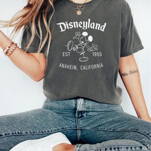 Puede incluir: Camiseta color pimienta con el logo de Disneyland, Mickey Mouse, el a&ntilde;o de fundaci&oacute;n 1955 y Anaheim, California. La camiseta se combina con vaqueros azules y zapatillas blancas. El estilo es informal y c&oacute;modo.