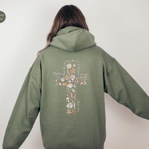 Puede incluir: Sudadera con capucha verde militar con un dise&ntilde;o de cruz en la espalda, adornada con conchas marinas y texto. La sudadera tiene capucha y mangas largas. El texto en la cruz dice "M&aacute;s poderoso que las olas del mar es su amor por ti".