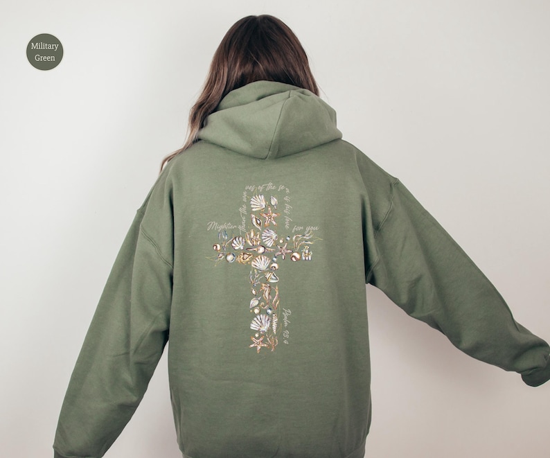 Puede incluir: Sudadera con capucha verde militar con un dise&ntilde;o de cruz en la espalda, adornada con conchas marinas y texto. La sudadera tiene capucha y mangas largas. El texto en la cruz dice "M&aacute;s poderoso que las olas del mar es su amor por ti".