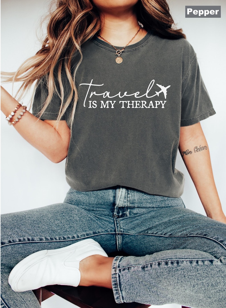 May include: A dark grey t-shirt with the words "Travel is my Therapy" in white script and a small airplane graphic. The shirt is worn with blue jeans and white trainers, creating a casual, travel-themed outfit.