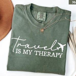 May include: A moss green t-shirt with the white text "Travel is my Therapy" and a plane graphic. A gold necklace with a pendant is on the shirt. A brown hat is visible in the upper left corner.