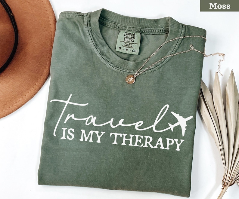 May include: A moss green t-shirt with the white text "Travel is my Therapy" and a plane graphic. A gold necklace with a pendant is on the shirt. A brown hat is visible in the upper left corner.