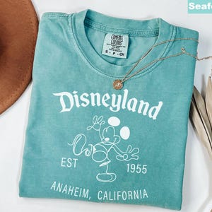 Puede incluir: Camiseta verde mar con la palabra "Disneyland" en escritura blanca, junto con un gr&aacute;fico de Mickey Mouse. La camiseta tambi&eacute;n presenta el texto "EST 1955" y "Anaheim, California". Un collar dorado con un colgante descansa sobre la camiseta.