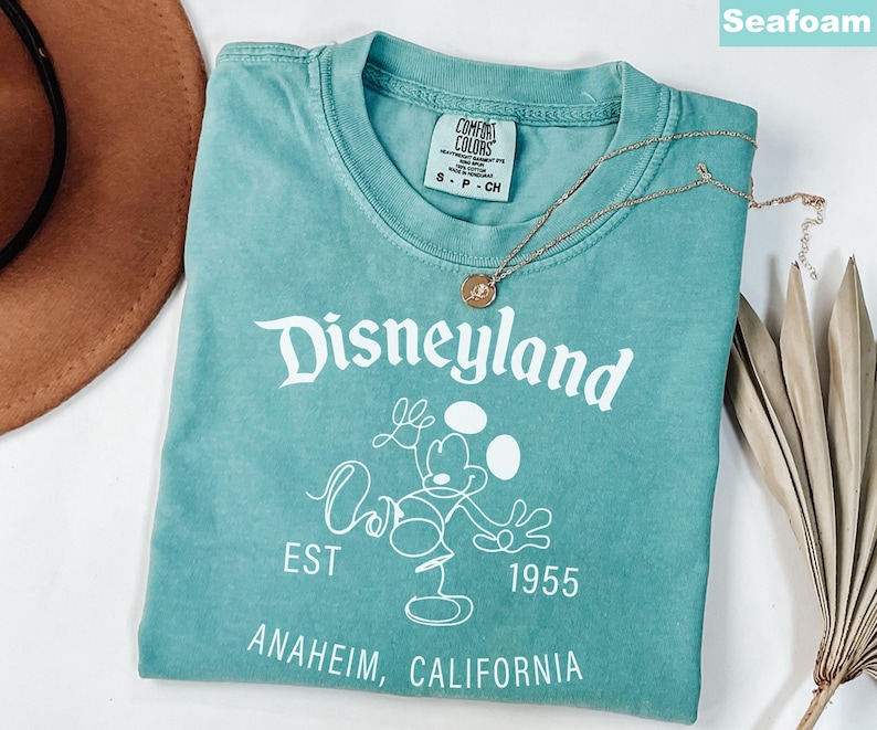 Puede incluir: Camiseta verde mar con la palabra "Disneyland" en escritura blanca, junto con un gr&aacute;fico de Mickey Mouse. La camiseta tambi&eacute;n presenta el texto "EST 1955" y "Anaheim, California". Un collar dorado con un colgante descansa sobre la camiseta.