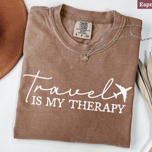May include: Espresso-coloured t-shirt with the words "Travel is my Therapy" in white script and a plane graphic. A silver necklace with a pendant rests on the shirt. The shirt is a Comfort Colors brand.