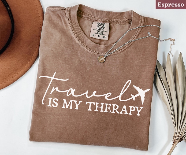 May include: Espresso-coloured t-shirt with the words "Travel is my Therapy" in white script and a plane graphic. A silver necklace with a pendant rests on the shirt. The shirt is a Comfort Colors brand.