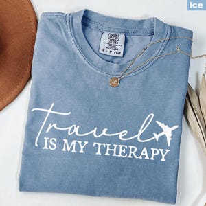 May include: Ice blue Comfort Colors t-shirt with the text "Travel is my Therapy" and an airplane graphic. A gold necklace with a pendant rests on the shirt. A brown hat and dried plant are in the background.