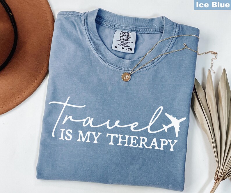 May include: Ice blue Comfort Colors t-shirt with the text "Travel is my Therapy" and an airplane graphic. A gold necklace with a pendant rests on the shirt. A brown hat and dried plant are in the background.