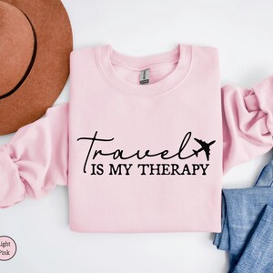 May include: A pale pink sweatshirt featuring the black text "Travel is my therapy" and an airplane graphic. A brown hat and blue jeans are also visible in the image.