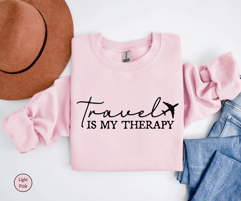 May include: A pale pink sweatshirt featuring the black text "Travel is my therapy" and an airplane graphic. A brown hat and blue jeans are also visible in the image.