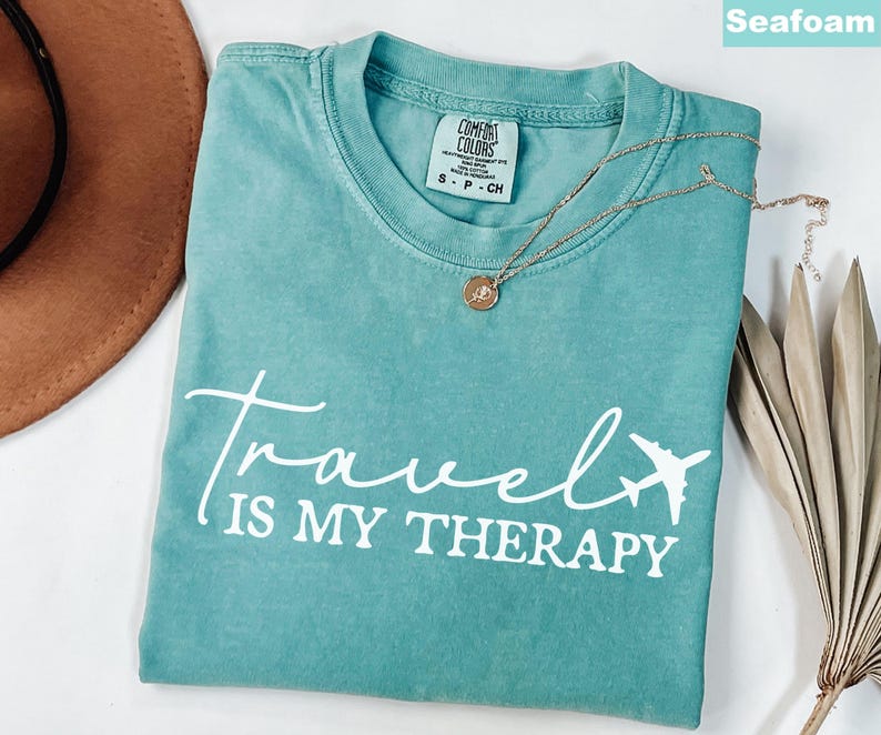 May include: Seafoam green t-shirt with the text "Travel is my Therapy" in white script and a white airplane graphic. A gold necklace with a pendant is on the shirt. A brown hat and dried plant are in the background.