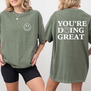 Puede incluir: Una camiseta verde musgo con una carita sonriente en la parte delantera y la frase "YOU'RE DOING GREAT" en la espalda. La camiseta tiene un corte holgado y mangas cortas. La modelo lleva un pantal&oacute;n corto negro.