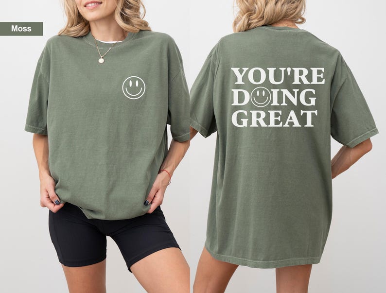 Puede incluir: Una camiseta verde musgo con una carita sonriente en la parte delantera y la frase "YOU'RE DOING GREAT" en la espalda. La camiseta tiene un corte holgado y mangas cortas. La modelo lleva un pantal&oacute;n corto negro.