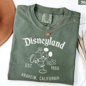 Puede incluir: Una camiseta verde musgo con el logotipo de Disneyland en blanco, con Mickey Mouse. La camiseta incluye el texto "EST 1955" y "ANAHEIM, CALIFORNIA". Tambi&eacute;n se ven un collar dorado y un sombrero marr&oacute;n.