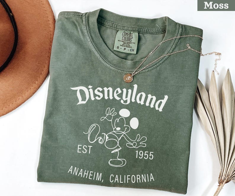 Puede incluir: Una camiseta verde musgo con el logotipo de Disneyland en blanco, con Mickey Mouse. La camiseta incluye el texto "EST 1955" y "ANAHEIM, CALIFORNIA". Tambi&eacute;n se ven un collar dorado y un sombrero marr&oacute;n.