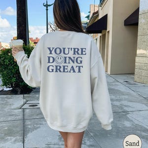 Puede incluir: Sudadera color arena con el texto azul "YOU'RE DOING GREAT" y un emoticono sonriente. La sudadera es de cuello redondo y parece estar hecha de un material suave. La palabra "Sand" est&aacute; en un c&iacute;rculo en la esquina inferior derecha.