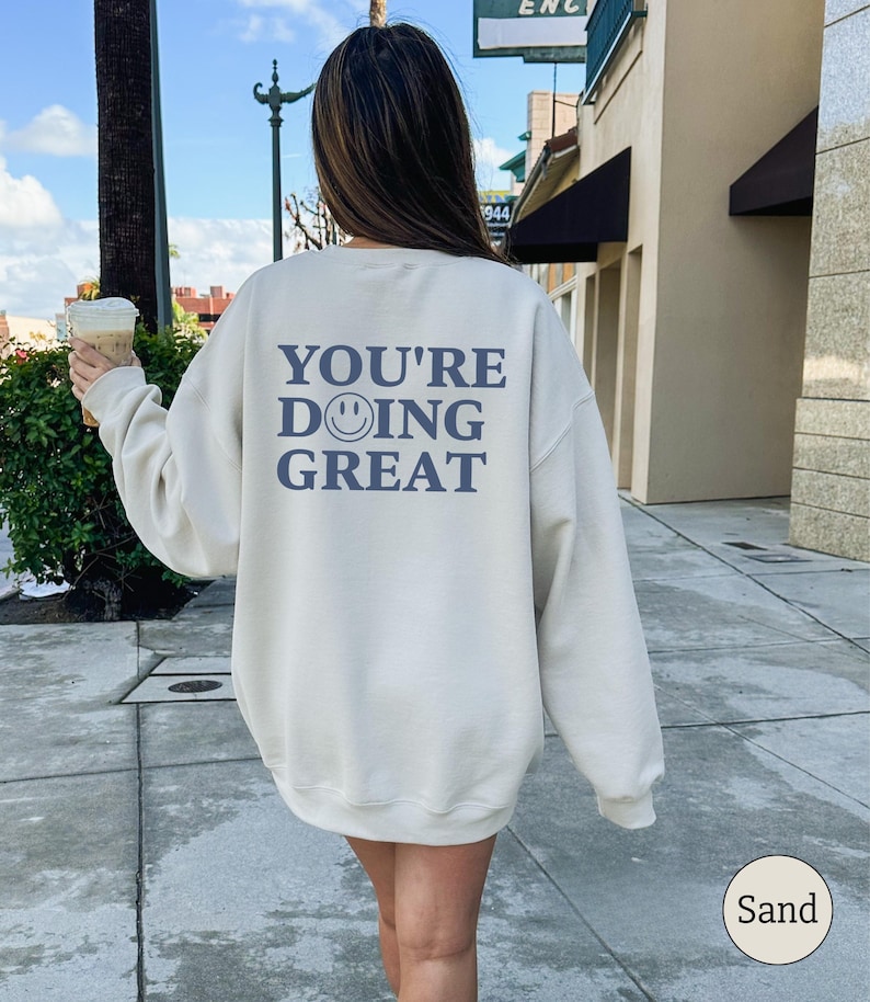 Puede incluir: Sudadera color arena con el texto azul "YOU'RE DOING GREAT" y un emoticono sonriente. La sudadera es de cuello redondo y parece estar hecha de un material suave. La palabra "Sand" est&aacute; en un c&iacute;rculo en la esquina inferior derecha.