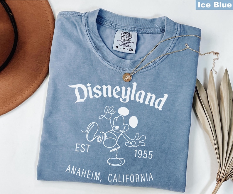Puede incluir: Camiseta azul hielo con el logotipo de Disneyland y un gr&aacute;fico de Mickey Mouse. La camiseta incluye el texto "EST 1955 ANAHEIM, CALIFORNIA". Un collar dorado est&aacute; sobre la camiseta.