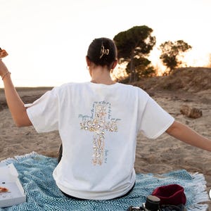 Puede incluir: Camiseta blanca con un dise&ntilde;o de cruz hecho de flores y texto en la espalda. La camiseta se usa en una playa con una manta azul y pizza. El texto en la camiseta dice "M&aacute;s poderoso que las olas del mar es su amor por ti."