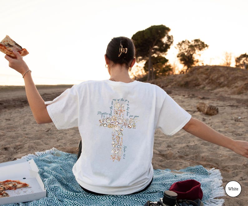 Puede incluir: Camiseta blanca con un dise&ntilde;o de cruz hecho de flores y texto en la espalda. La camiseta se usa en una playa con una manta azul y pizza. El texto en la camiseta dice "M&aacute;s poderoso que las olas del mar es su amor por ti."