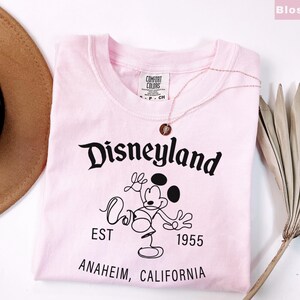 Puede incluir: Una camiseta rosa claro con la palabra "Disneyland" en escritura negra, junto con un dibujo de Mickey Mouse. La camiseta tambi&eacute;n presenta el texto "EST 1955" y "ANAHEIM, CALIFORNIA". Un collar dorado con un colgante descansa sobre la camiseta.