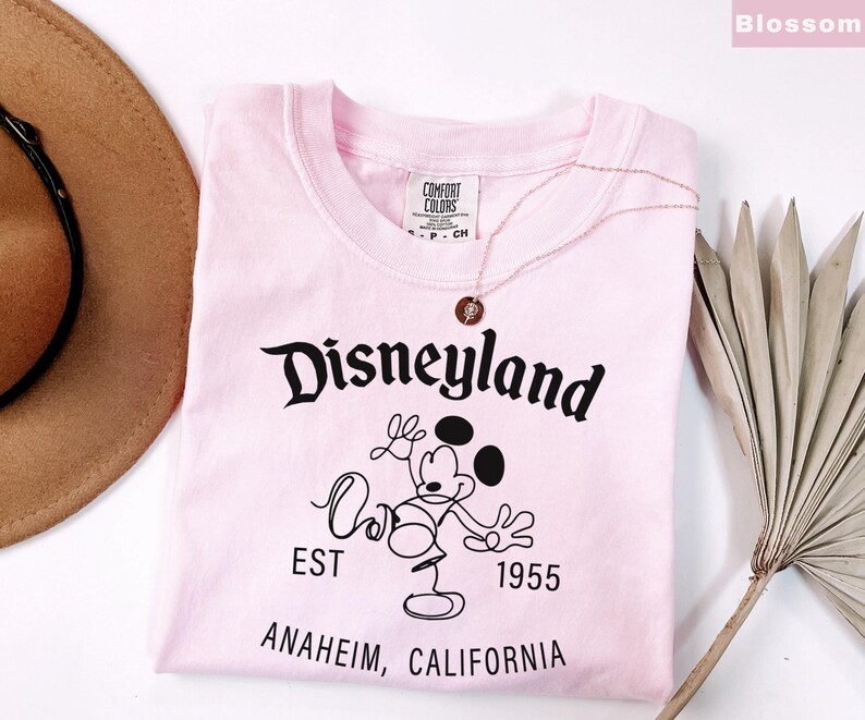 Puede incluir: Una camiseta rosa claro con la palabra "Disneyland" en escritura negra, junto con un dibujo de Mickey Mouse. La camiseta tambi&eacute;n presenta el texto "EST 1955" y "ANAHEIM, CALIFORNIA". Un collar dorado con un colgante descansa sobre la camiseta.