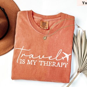 May include: A yam-coloured t-shirt with the text "Travel is my Therapy" in white script and a small airplane graphic. The shirt has a crew neck and a small tag. A gold necklace and a brown hat are also in the image.