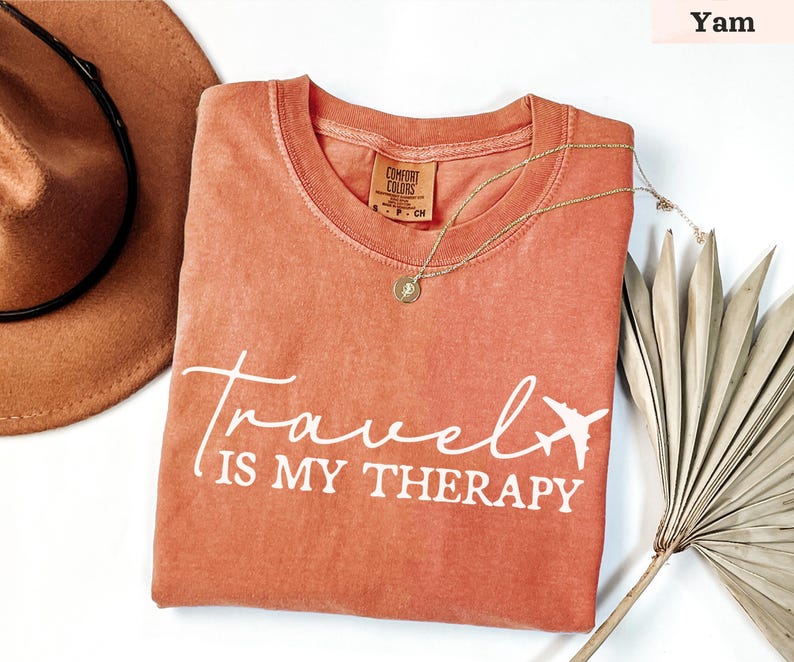 May include: A yam-coloured t-shirt with the text "Travel is my Therapy" in white script and a small airplane graphic. The shirt has a crew neck and a small tag. A gold necklace and a brown hat are also in the image.