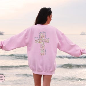 Puede incluir: Sudadera rosa claro con un dise&ntilde;o de cruz en la espalda. La cruz est&aacute; hecha de conchas marinas y elementos florales, con texto incorporado en el dise&ntilde;o. La sudadera es de color rosa claro liso.